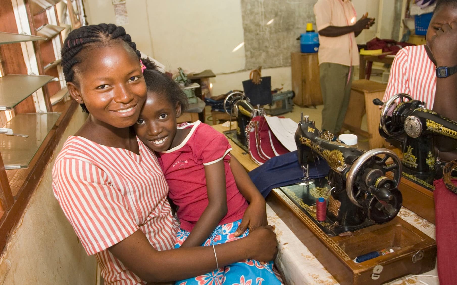 A teenage girl holds a young girl in her lap while they are sewing on the sewing machine