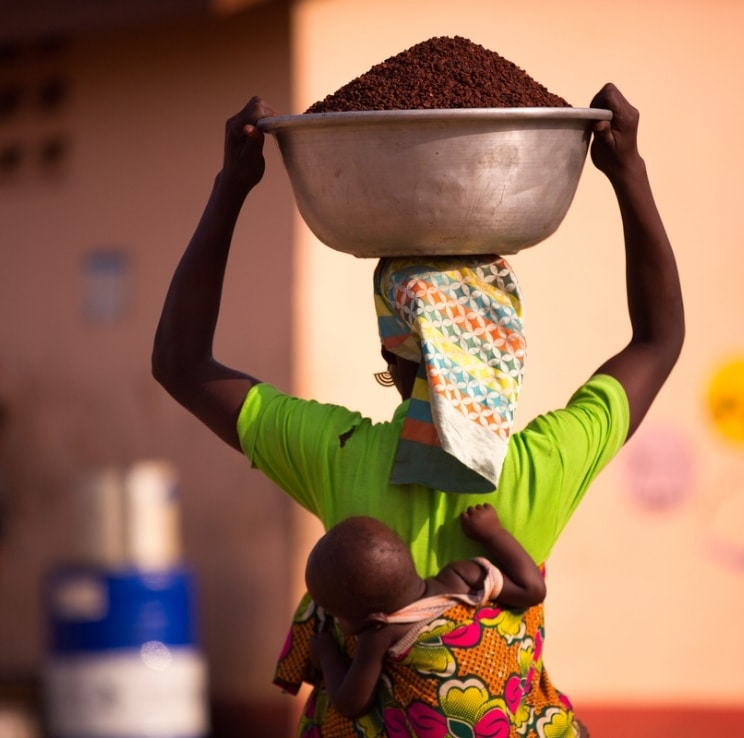 A woman carrying a bowl with grains in it while carrying her baby on her back