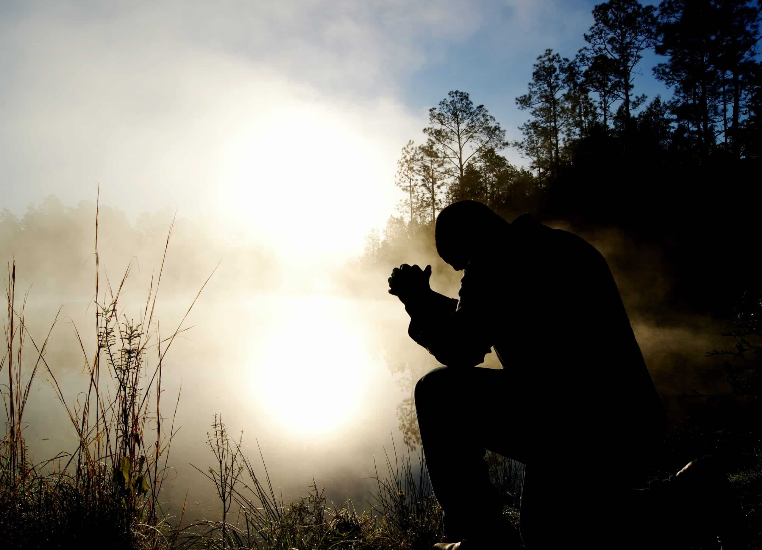 The silhouette of a man is kneeling in prayer in a field by a pond lit by sunlight. Demons Still Flee at the Mention of Jesus’ Name