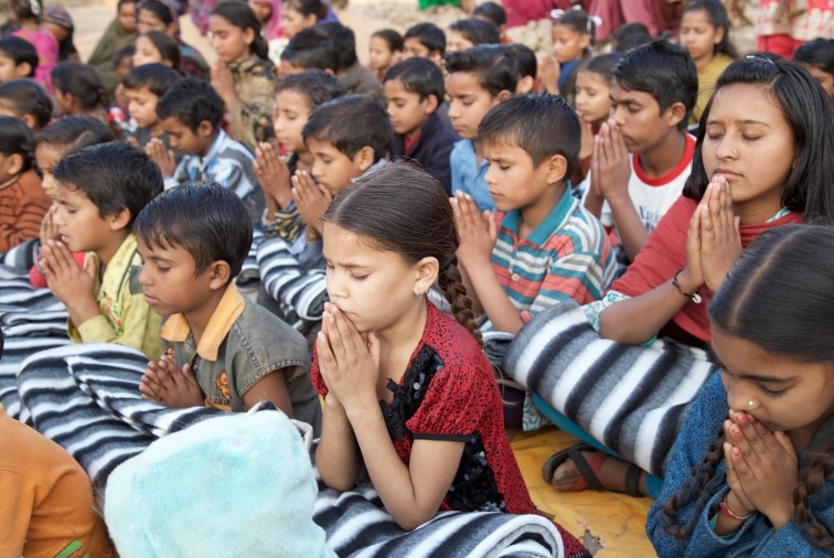 little kids siting on the ground outside and praying with their hands folded and eyes closed