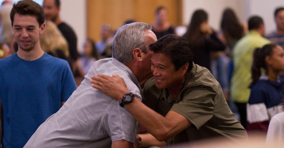 Two smiling men shake hands and embrace at a church missions conference with other attendees in the background. How ANM Helps Churches Engage in Global Missions