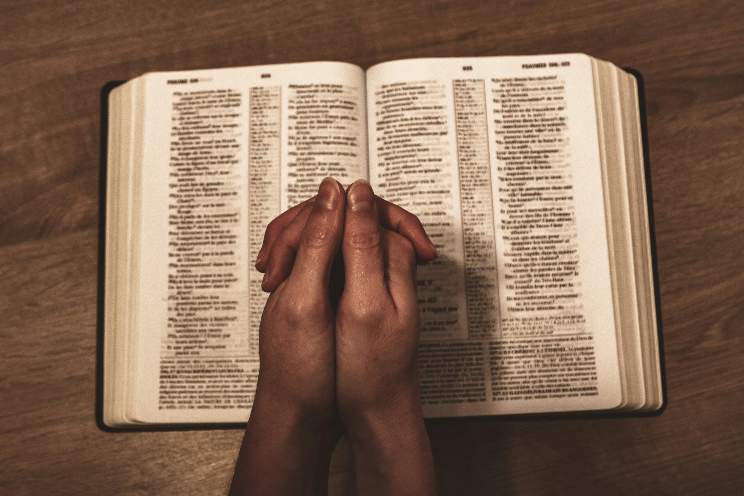 Hands clasped in prayer rest on an open Bible, lying on a wooden table. The scene conveys a sense of peace, faith, and contemplation.
